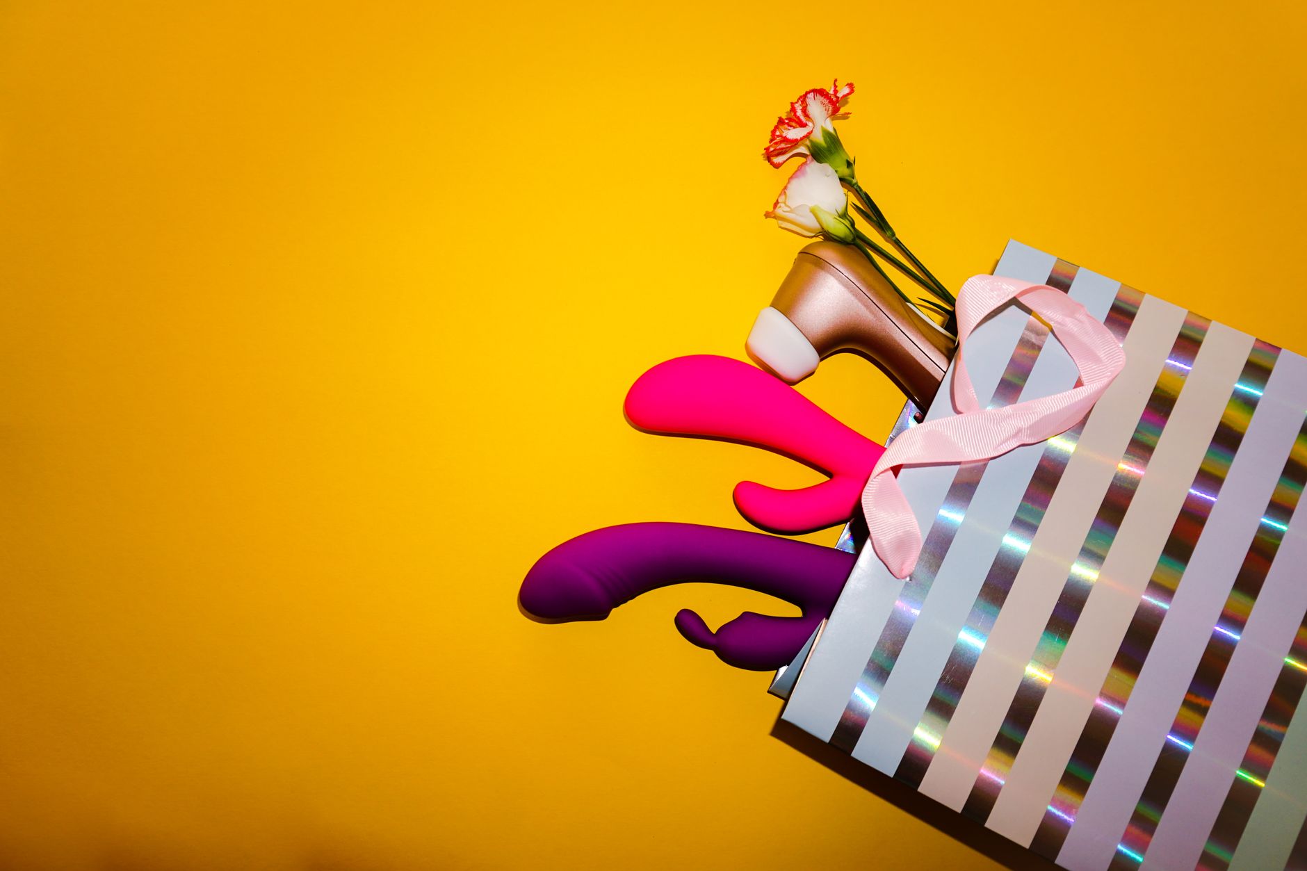 Colorful vibrators with flowers arranged in a holographic gift bag against a bold yellow background.